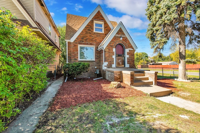 tudor house featuring roof with shingles, a front yard, fence, and brick siding