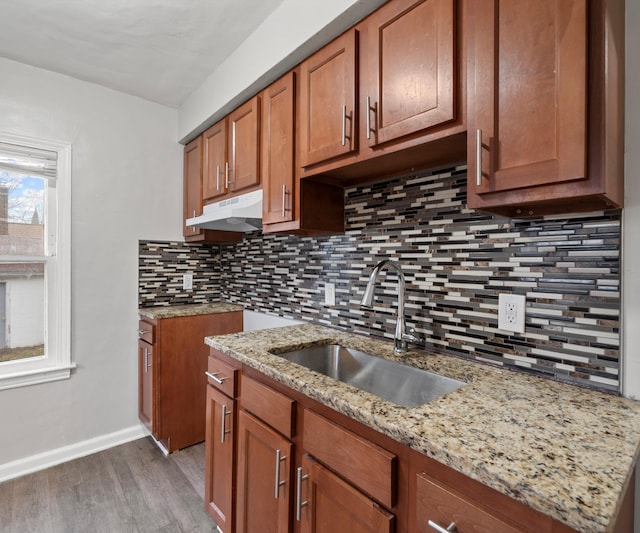 kitchen featuring brown cabinetry, wood finished floors, light stone countertops, under cabinet range hood, and a sink