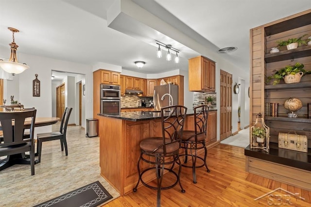 kitchen with a breakfast bar area, brown cabinets, stainless steel appliances, under cabinet range hood, and backsplash