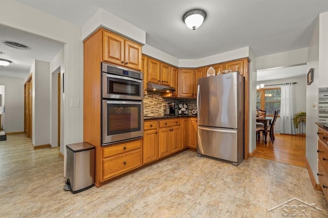 kitchen featuring brown cabinetry, dark countertops, appliances with stainless steel finishes, under cabinet range hood, and backsplash