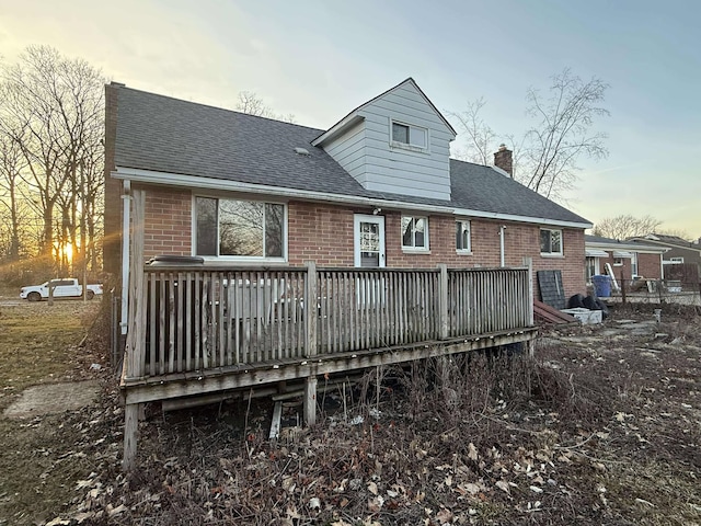 back of house at dusk with brick siding, roof with shingles, a deck, and a chimney