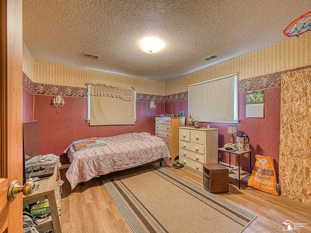 bedroom with a textured ceiling, wood finished floors, visible vents, and wallpapered walls