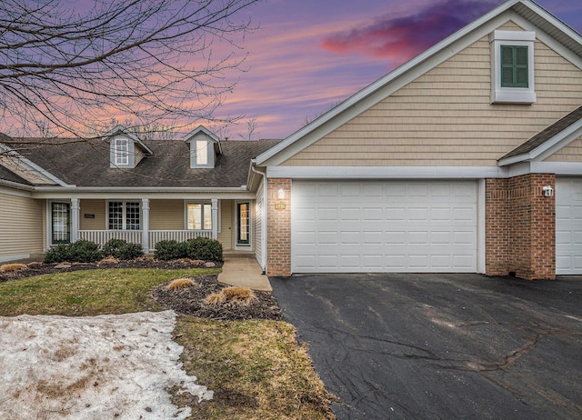 cape cod-style house with a garage, a shingled roof, aphalt driveway, covered porch, and brick siding