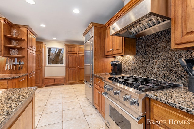 kitchen featuring premium appliances, wall chimney range hood, backsplash, dark stone counters, and brown cabinetry