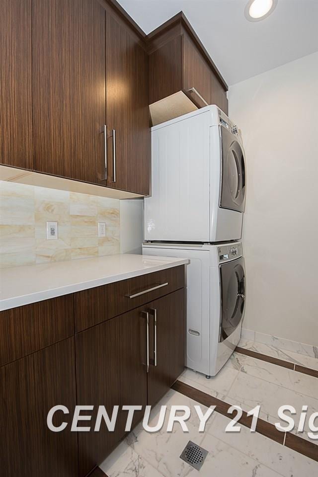 clothes washing area featuring marble finish floor, stacked washing maching and dryer, cabinet space, and baseboards