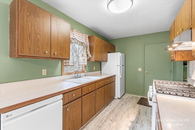 kitchen with white appliances, brown cabinetry, light countertops, under cabinet range hood, and a sink