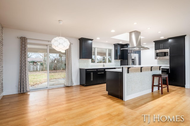 kitchen with island range hood, stainless steel appliances, a kitchen breakfast bar, light countertops, and dark cabinetry