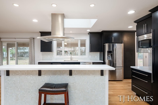 kitchen featuring island exhaust hood, stainless steel appliances, light countertops, dark cabinetry, and a kitchen bar