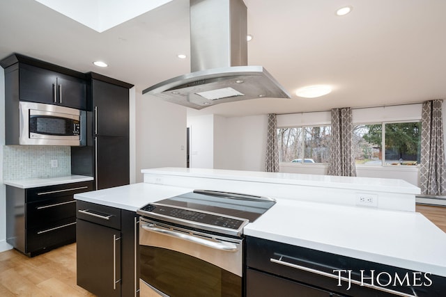 kitchen featuring island range hood, appliances with stainless steel finishes, light countertops, dark cabinetry, and light wood-style floors