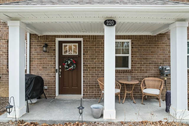 doorway to property featuring a porch, roof with shingles, and brick siding