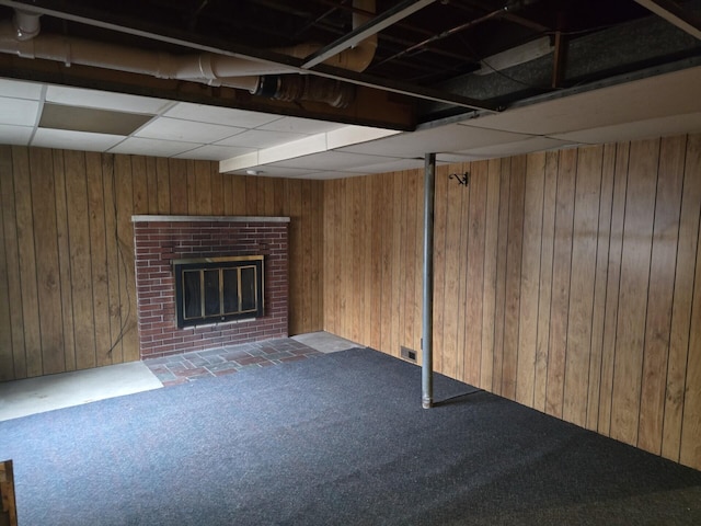 finished basement featuring wooden walls, visible vents, a drop ceiling, carpet flooring, and a brick fireplace