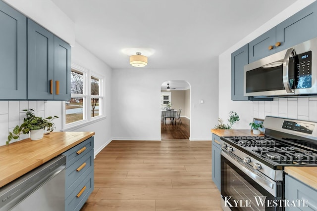 kitchen with arched walkways, decorative backsplash, appliances with stainless steel finishes, light wood-style floors, and butcher block countertops