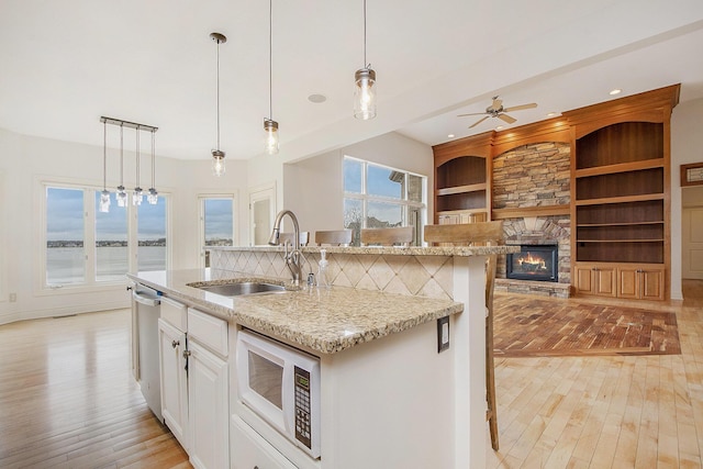 kitchen with dishwasher, white microwave, a stone fireplace, light wood-type flooring, and a sink