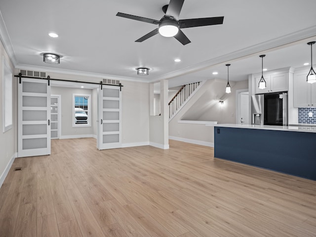 unfurnished living room featuring crown molding, light wood-style flooring, stairway, a barn door, and a ceiling fan