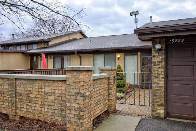 doorway to property featuring brick siding, an attached garage, a shingled roof, and fence