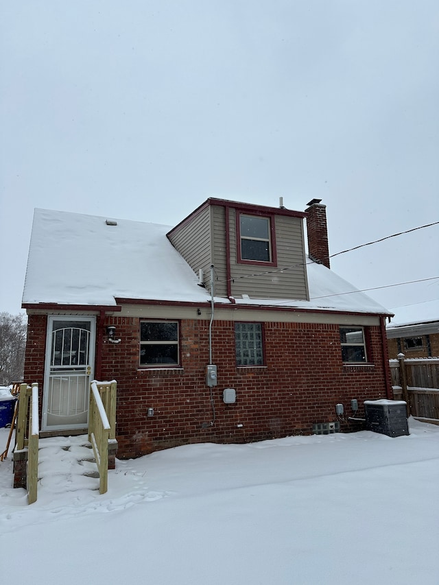 snow covered property with central AC, brick siding, and a chimney