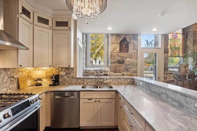 kitchen with stainless steel appliances, a sink, wall chimney range hood, backsplash, and light stone countertops