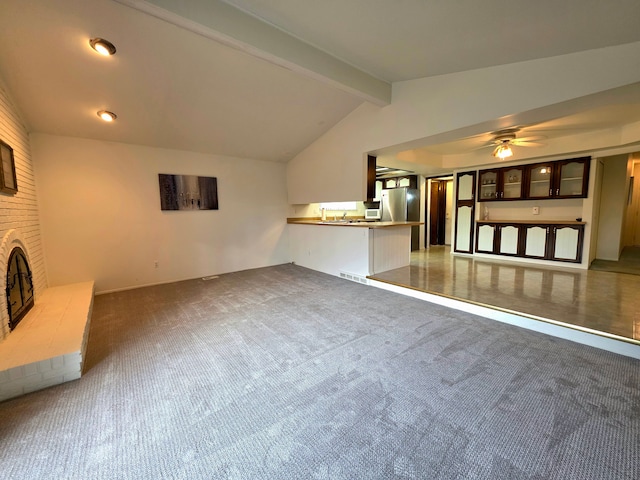 unfurnished living room with visible vents, a ceiling fan, vaulted ceiling with beams, a brick fireplace, and dark carpet