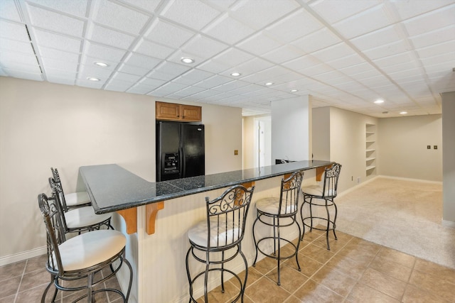 kitchen with brown cabinetry, baseboards, black fridge with ice dispenser, light carpet, and a kitchen bar