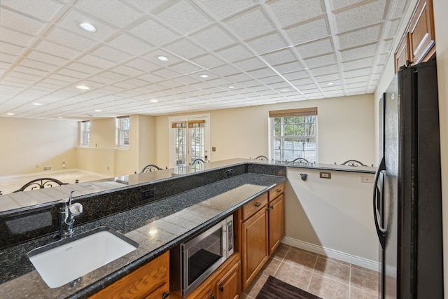 kitchen with a sink, stainless steel microwave, dark stone countertops, freestanding refrigerator, and brown cabinetry