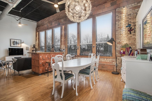 dining space featuring a towering ceiling, light wood finished floors, brick wall, and ceiling fan with notable chandelier