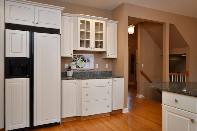 kitchen with white cabinets, light wood-style flooring, glass insert cabinets, dark stone countertops, and paneled built in refrigerator