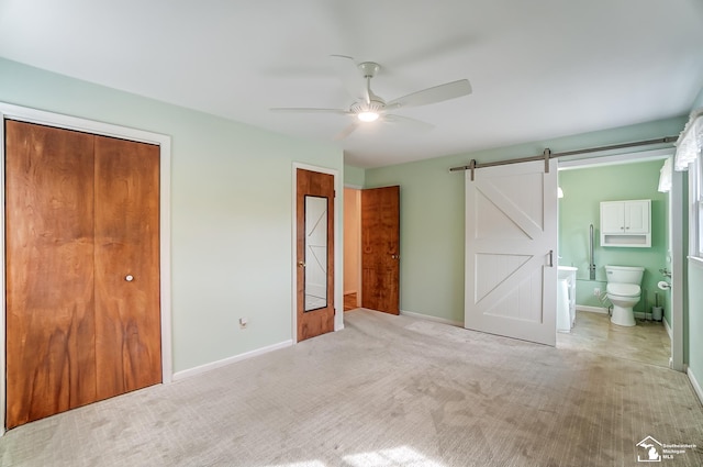 unfurnished bedroom with ensuite bathroom, a ceiling fan, a barn door, carpet flooring, and baseboards