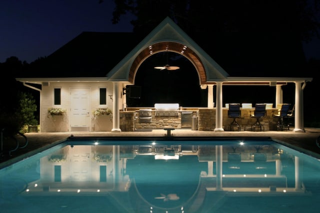 pool at night featuring a community pool, outdoor wet bar, a patio, and area for grilling