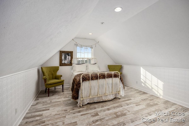 bedroom featuring vaulted ceiling, a textured ceiling, wood finished floors, baseboards, and wallpapered walls