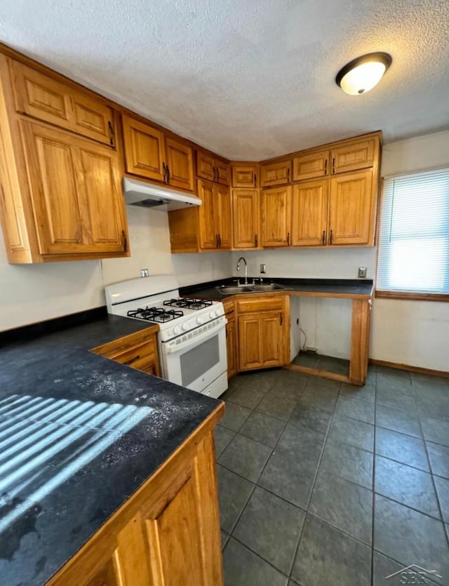 kitchen featuring under cabinet range hood, a sink, white gas range oven, brown cabinetry, and dark countertops