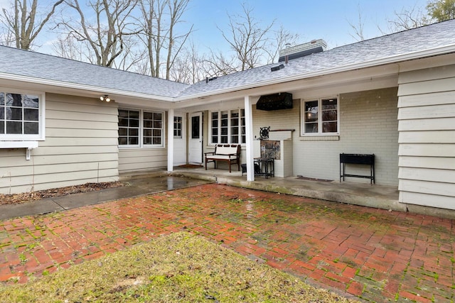 entrance to property featuring a shingled roof, a chimney, a patio, and brick siding