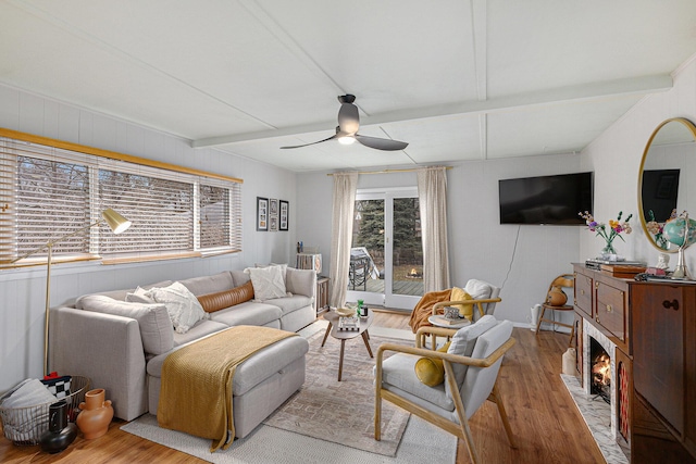 living area featuring a fireplace with flush hearth, beamed ceiling, wood finished floors, and a ceiling fan