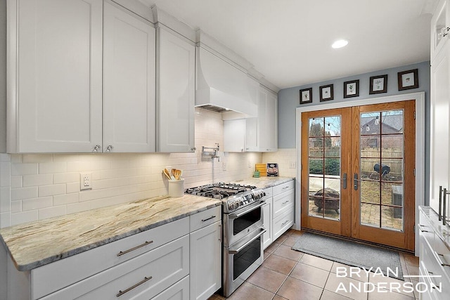 kitchen with light tile patterned floors, light stone counters, white cabinets, double oven range, and decorative backsplash