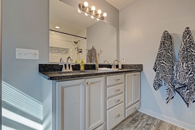 bathroom with wood finish floors, double vanity, a sink, tiled shower, and baseboards