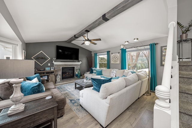 living area featuring a glass covered fireplace, lofted ceiling with beams, light wood-style flooring, ceiling fan, and stairway