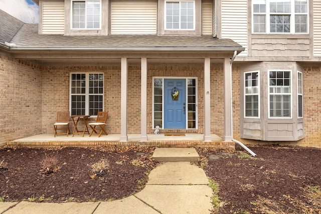 view of exterior entry with covered porch, roof with shingles, and brick siding