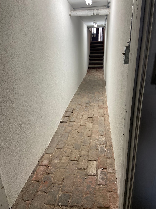 hallway featuring a textured wall, stairway, and brick floor