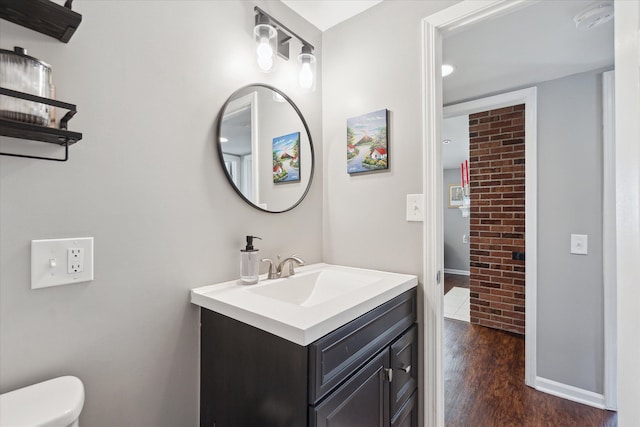 half bath with toilet, wood finished floors, brick wall, baseboards, and vanity