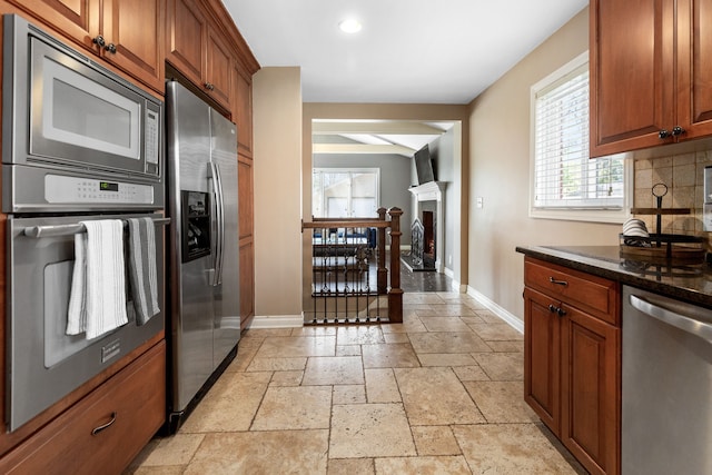 kitchen with brown cabinets, a fireplace with raised hearth, stone tile floors, stainless steel appliances, and baseboards