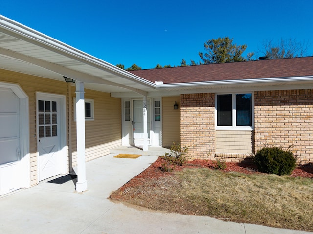 doorway to property featuring covered porch, brick siding, and roof with shingles