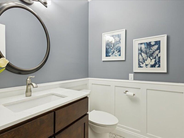 half bath with toilet, vanity, a decorative wall, and wainscoting