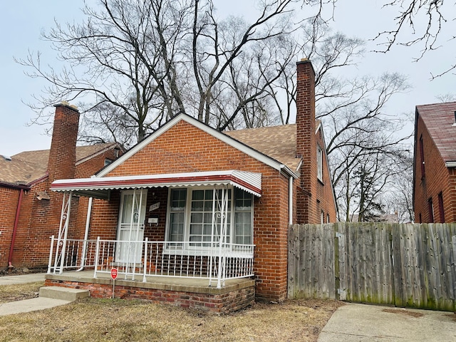 bungalow-style home featuring a porch, a shingled roof, brick siding, fence, and a chimney