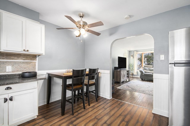 dining area with arched walkways, a wainscoted wall, dark wood-style floors, and a ceiling fan