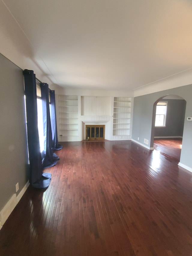 unfurnished living room with dark wood-style floors, built in shelves, arched walkways, a glass covered fireplace, and baseboards