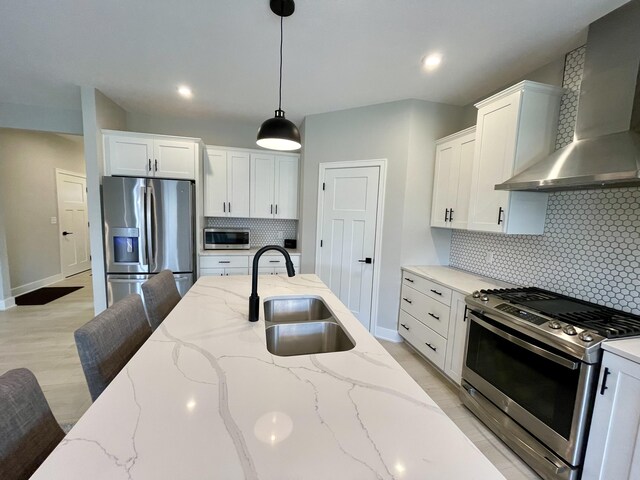 kitchen featuring a sink, white cabinets, appliances with stainless steel finishes, wall chimney range hood, and decorative light fixtures