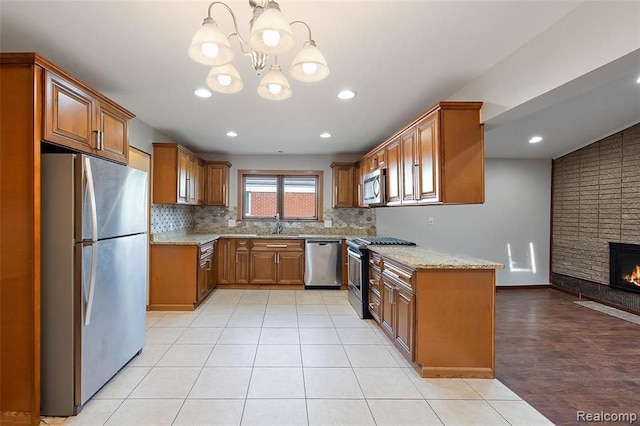 kitchen featuring a fireplace, appliances with stainless steel finishes, backsplash, brown cabinets, and light stone countertops