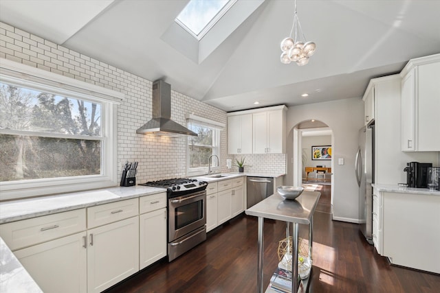 kitchen with a sink, stainless steel appliances, arched walkways, vaulted ceiling with skylight, and wall chimney exhaust hood