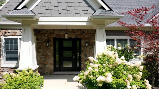 doorway to property featuring stone siding and roof with shingles