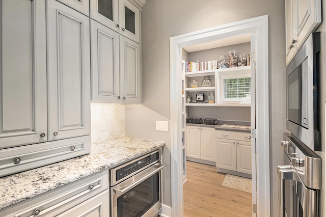 kitchen featuring light stone counters, light wood-style flooring, stainless steel appliances, backsplash, and glass insert cabinets