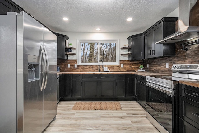 kitchen with dark cabinetry, open shelves, a sink, stainless steel appliances, and wall chimney range hood